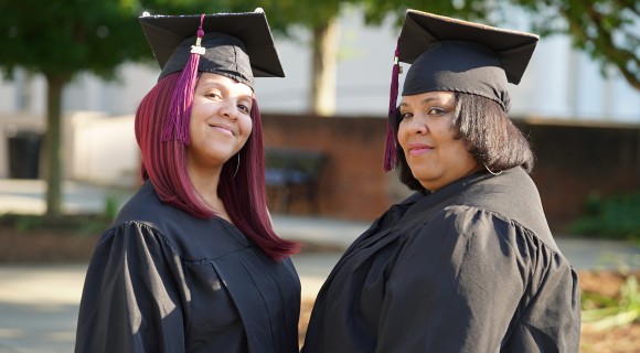 Two graduates in regalia stand on Mitchell's Statesville campus.